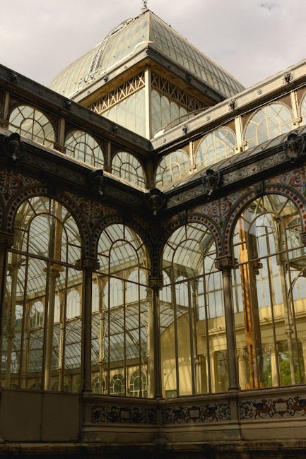 Close-up view of an ornate glass conservatory with intricate black metal framing and decorative details. The structure features large, arched glass panels and a prominent upper section with additional