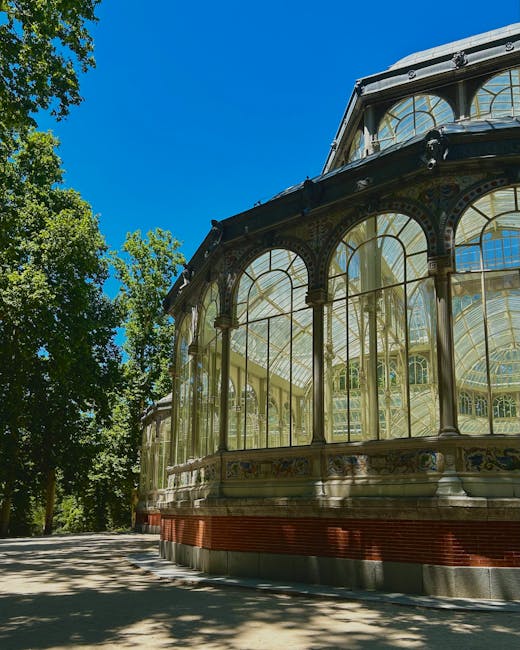 Close-up view of an ornate glass conservatory with intricate black metal framing and decorative details. The structure features large, arched glass panels and a prominent upper section with additional