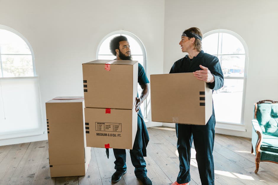 A young woman with curly black hair and casual clothing, including a white T-shirt and blue jeans, sitting on a wooden floor surrounded by numerous cardboard boxes of various sizes, some sealed with r